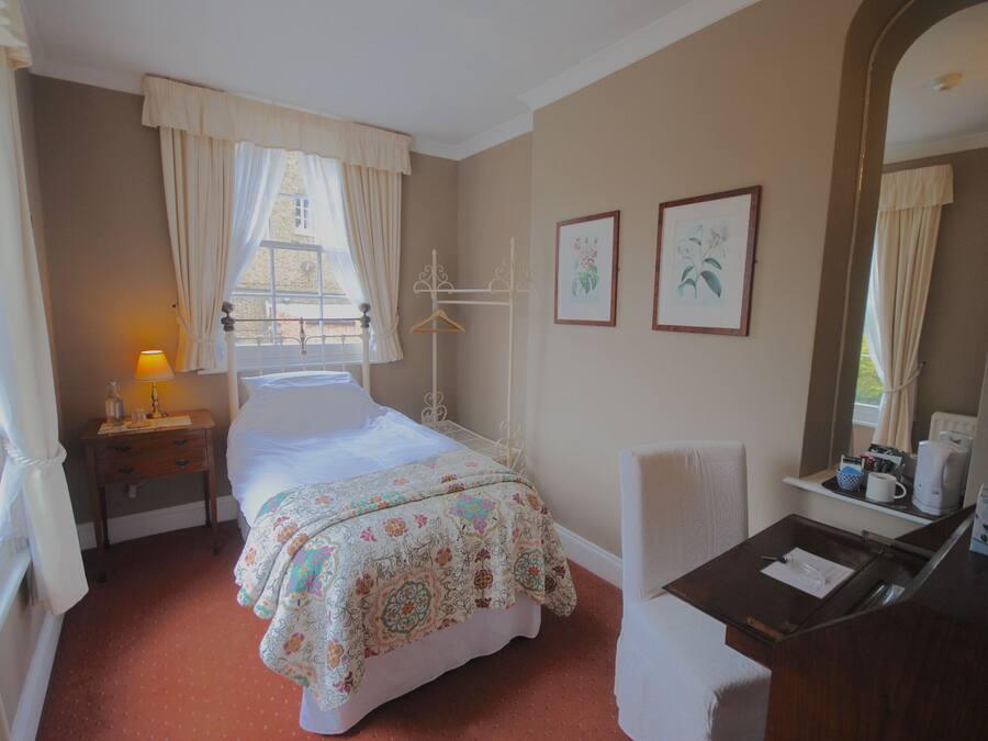 A hotel room with a bed, a desk with a chair, a window with curtains, a bedside table with a lamp, and framed pictures on the wall.