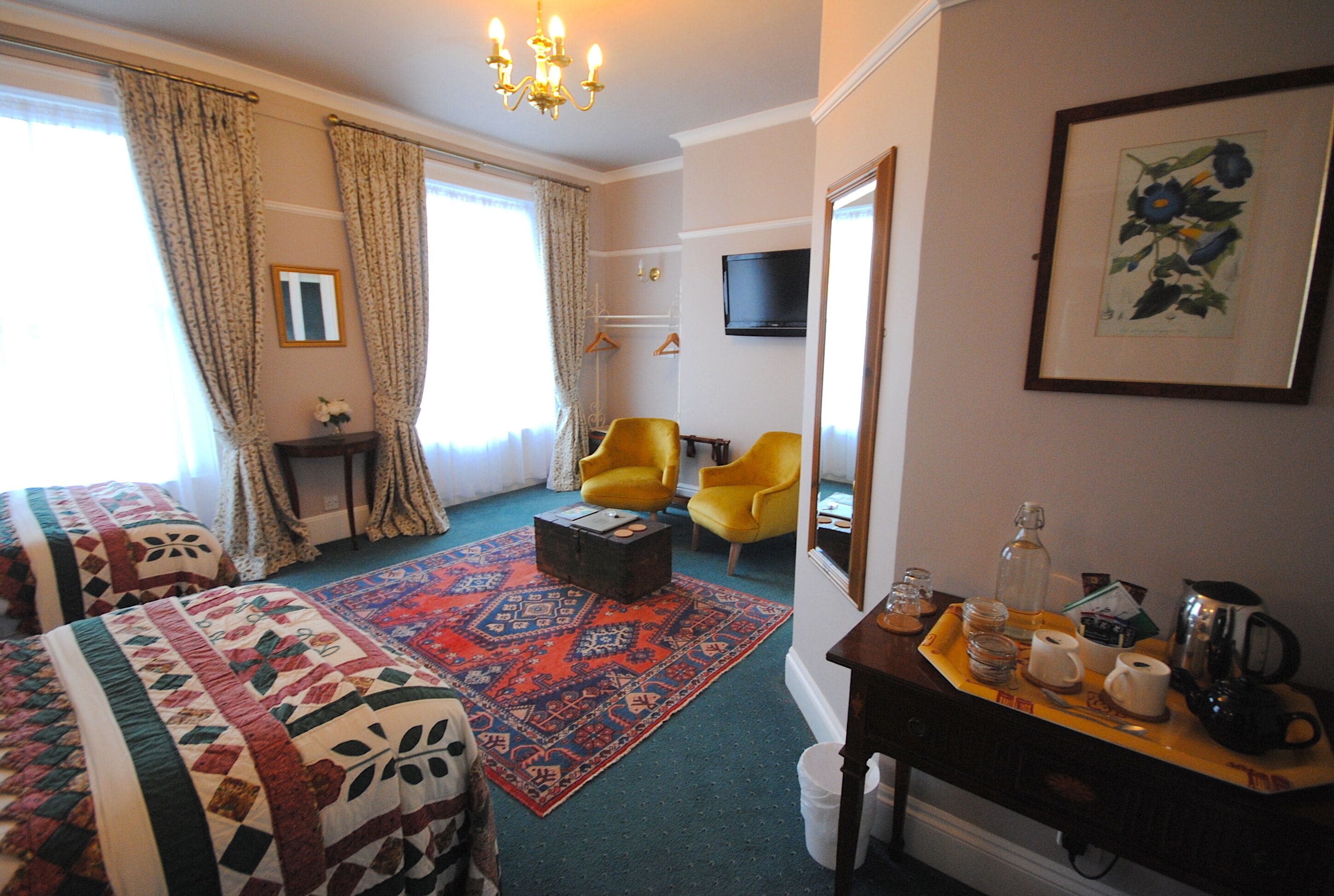 Une chambre d’hôtel avec deux lits, une grande fenêtre avec des rideaux, un téléviseur, un tableau encadré et une petite table sur laquelle se trouve un plateau avec divers objets.