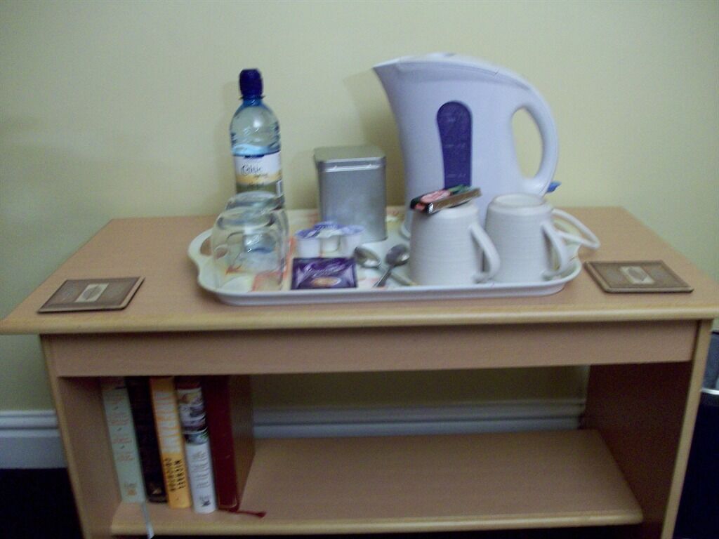 Una mesita con una botella de agua, una tetera, una taza, un pequeño recipiente y una bandeja con un teléfono y un libro.