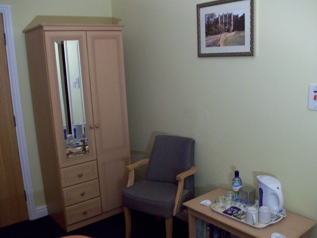 A room with a wooden wardrobe, a grey armchair, a small table with a kettle, cups, and a tray of snacks.