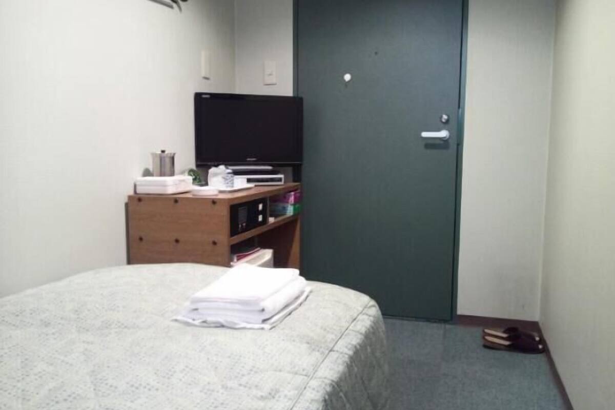 Um quarto pequeno com uma cama, uma cômoda de madeira, uma televisão e uma porta.