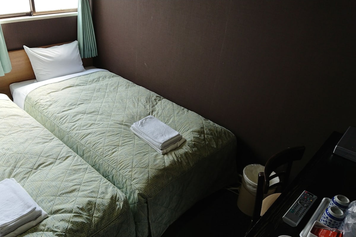 Quarto de hotel com duas camas, cada uma com colcha verde acolchoada e travesseiro branco. Há uma pequena mesa com um controle remoto, um pote e um tabuleiro com objetos.