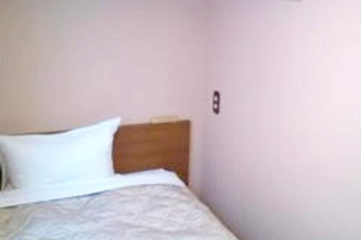 Uma cama de solteiro com cabeceira de madeira num quarto com paredes simples.