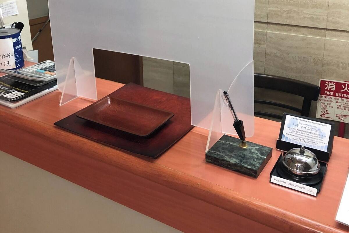 Uma mesa de recepção com monitor de computador, porta-canetas, uma pequena caixa e extintor de incêndio.