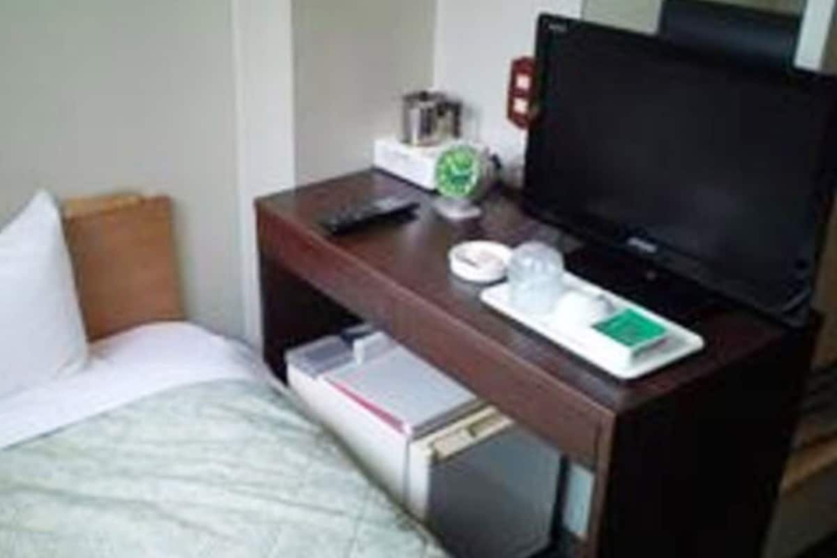 Quarto de hotel com uma cama, uma mesa de cabeceira de madeira com uma televisão, um controle remoto e uma pequena mesa com uma xícara e uma caixa de lenços.
