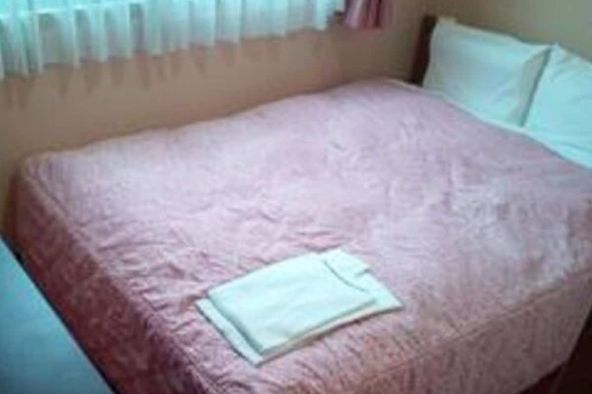Uma cama de solteiro com um cobertor rosa e um travesseiro branco em um quarto com uma janela coberta por cortinas finas.