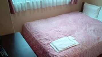 A single bed with a pink blanket and a white pillow in a room with a window covered by sheer curtains.