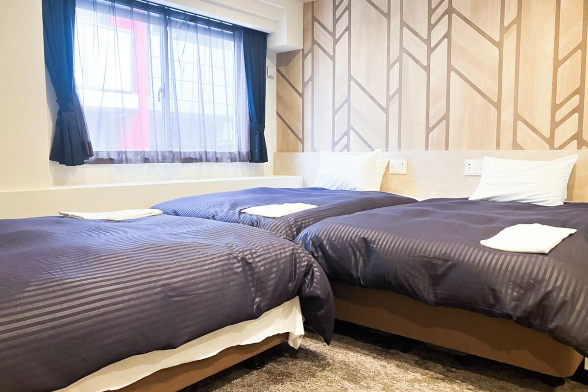 A hotel room with two beds, a window with sheer curtains, a geometric patterned wall, and a carpeted floor.