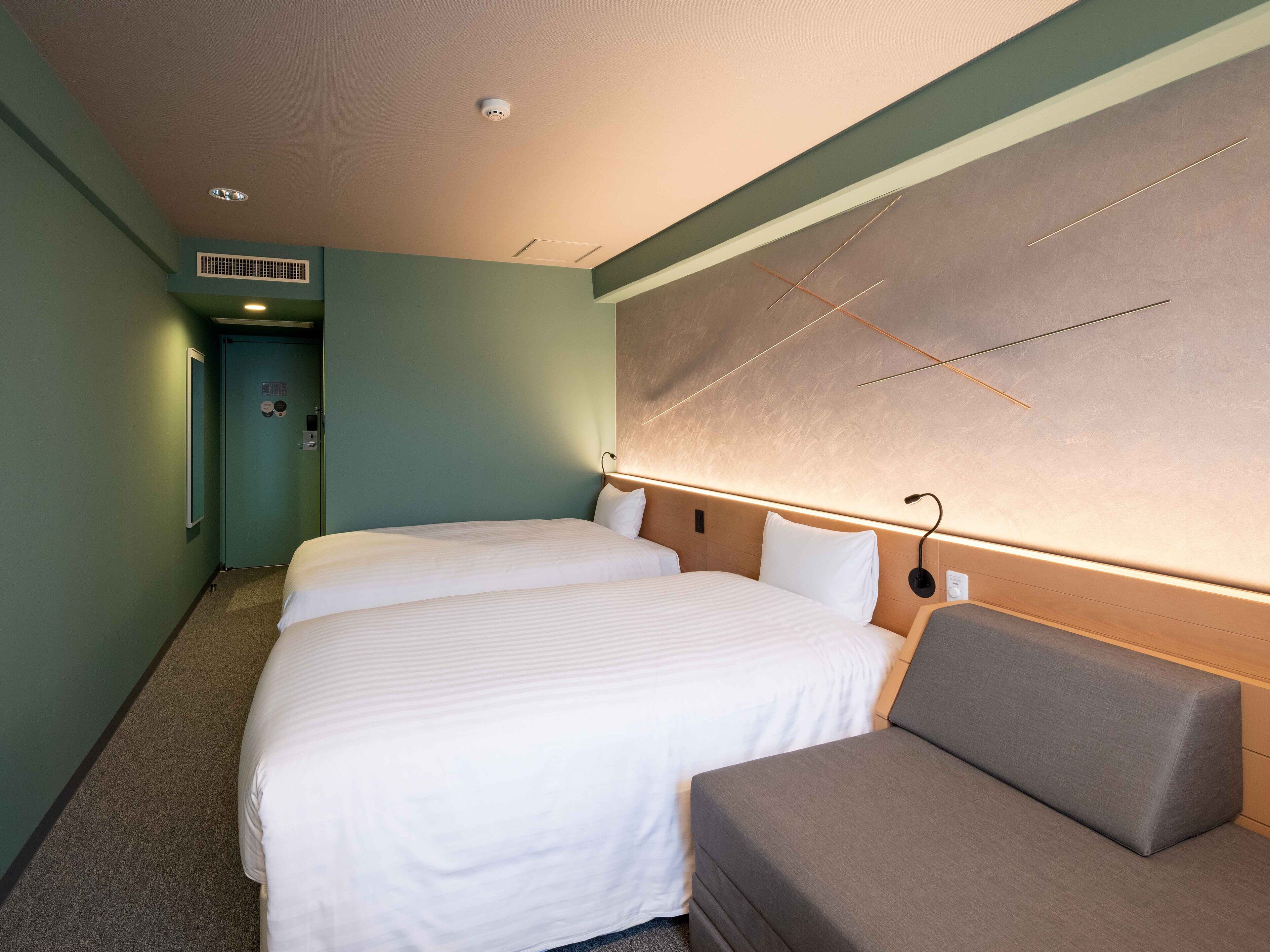 A hotel room with two beds, a sofa, a wall-mounted light, and a door with a vent.