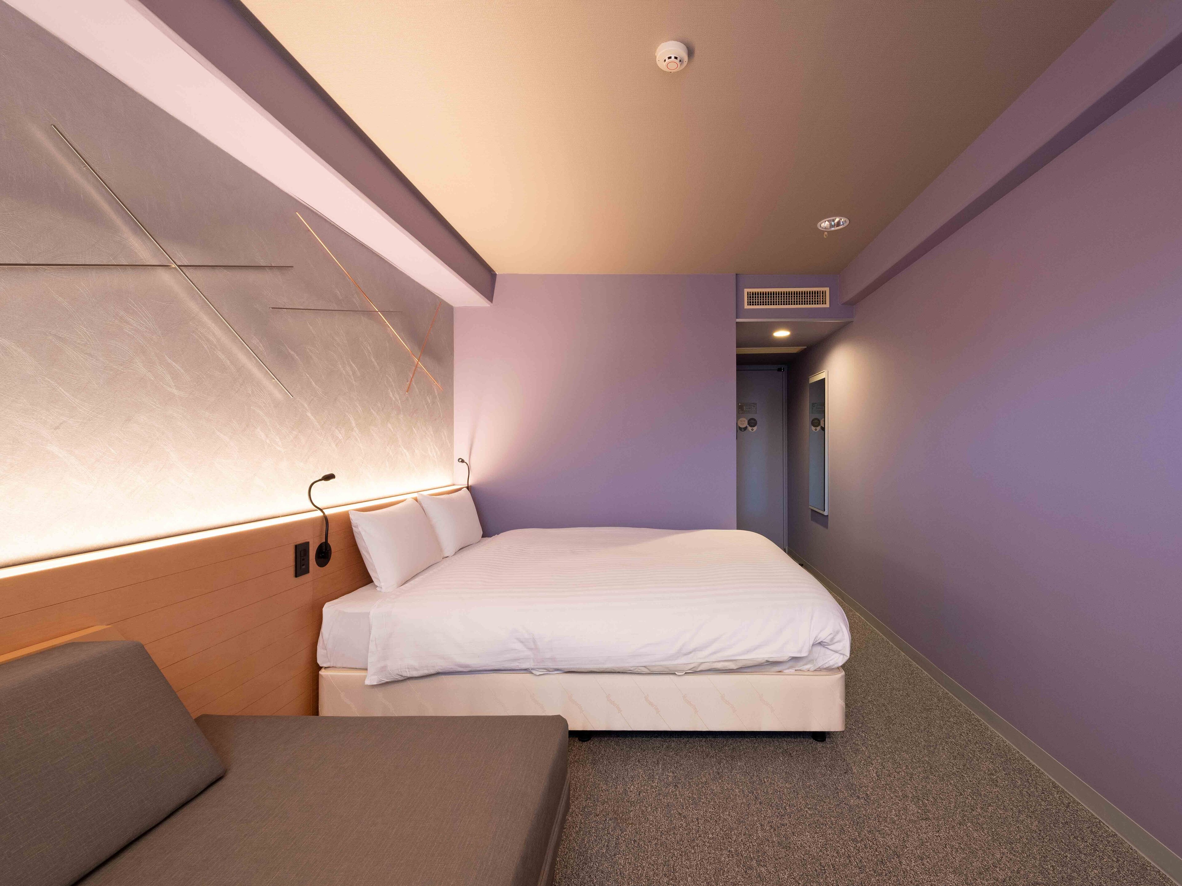 A modern hotel room with a bed, a sofa, a wall-mounted light, and a door.