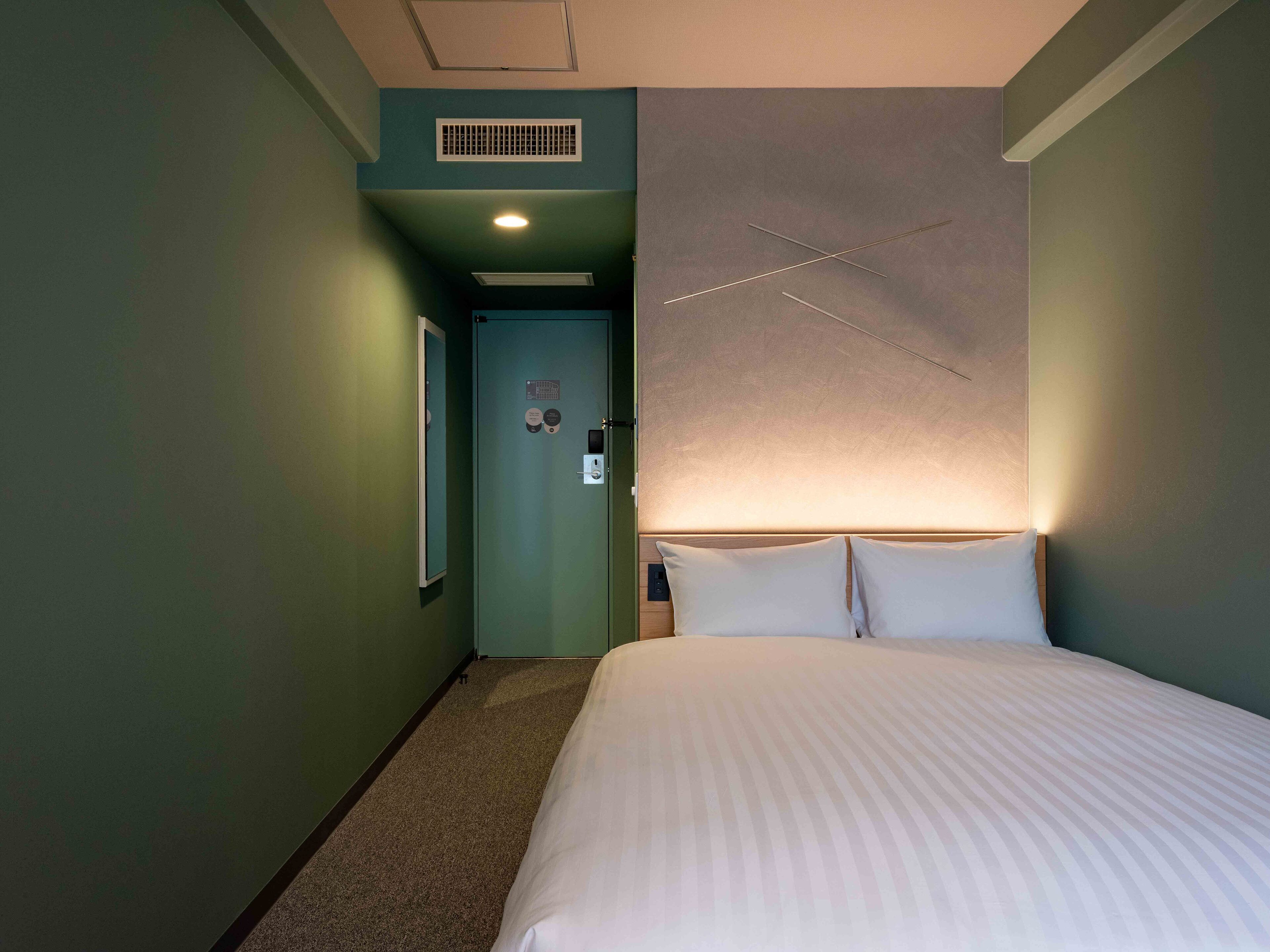 A hotel room with a bed, a nightstand, a wall-mounted light fixture, and a door leading to another room.
