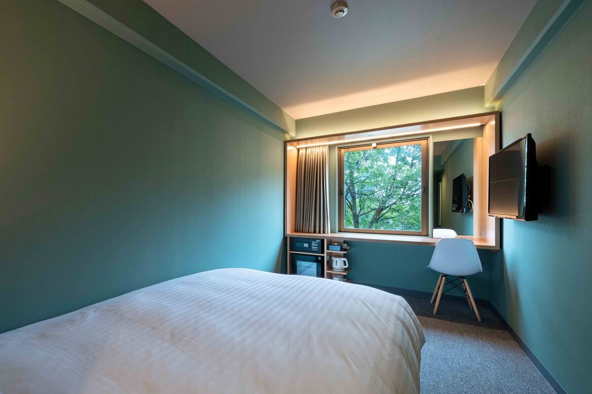Habitación de hotel con cama, escritorio con silla, televisor montado en la pared y ventana con vista a árboles.