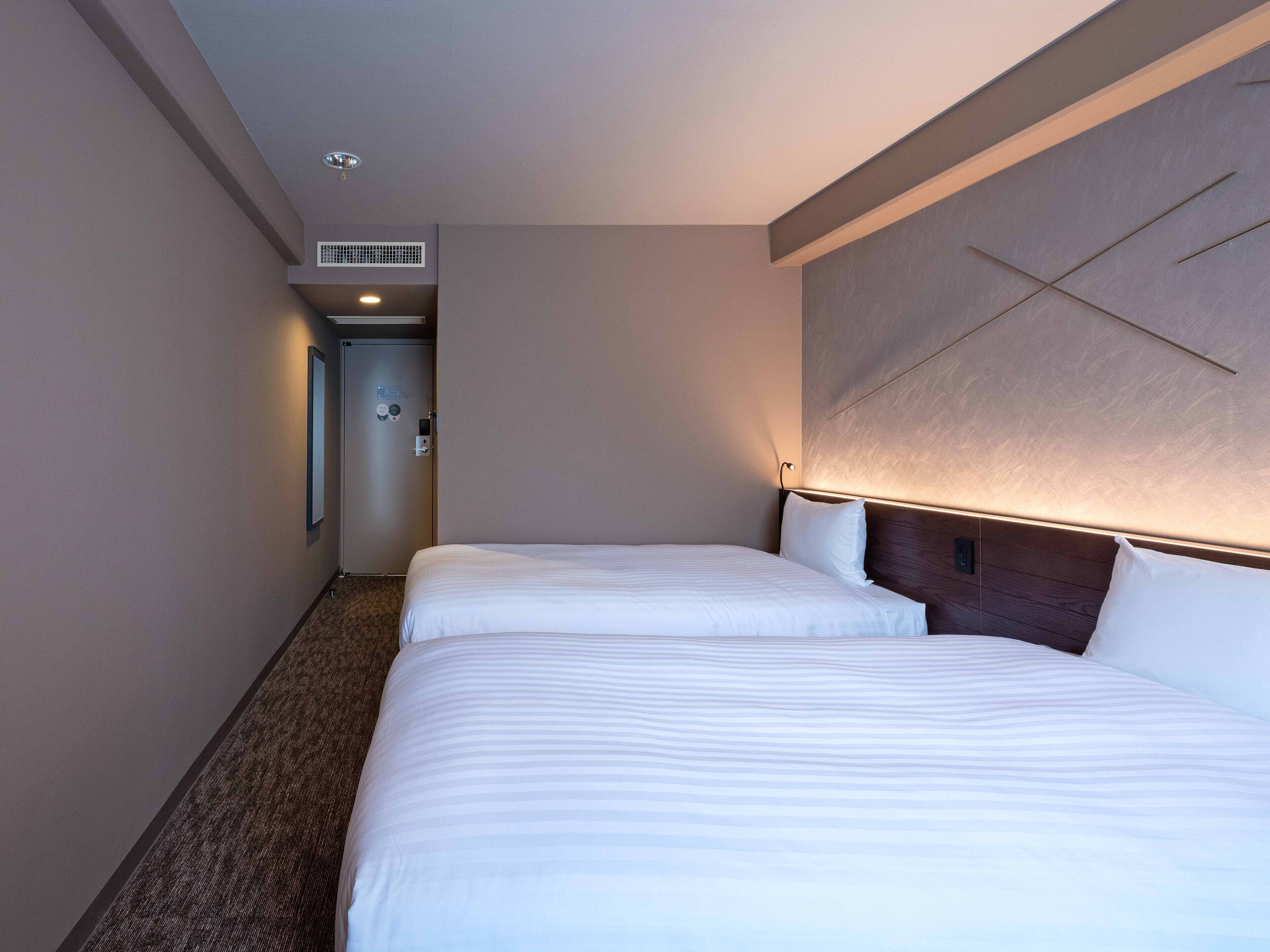 A hotel room with two beds, a wooden headboard, a carpeted floor, and a door leading to a hallway.