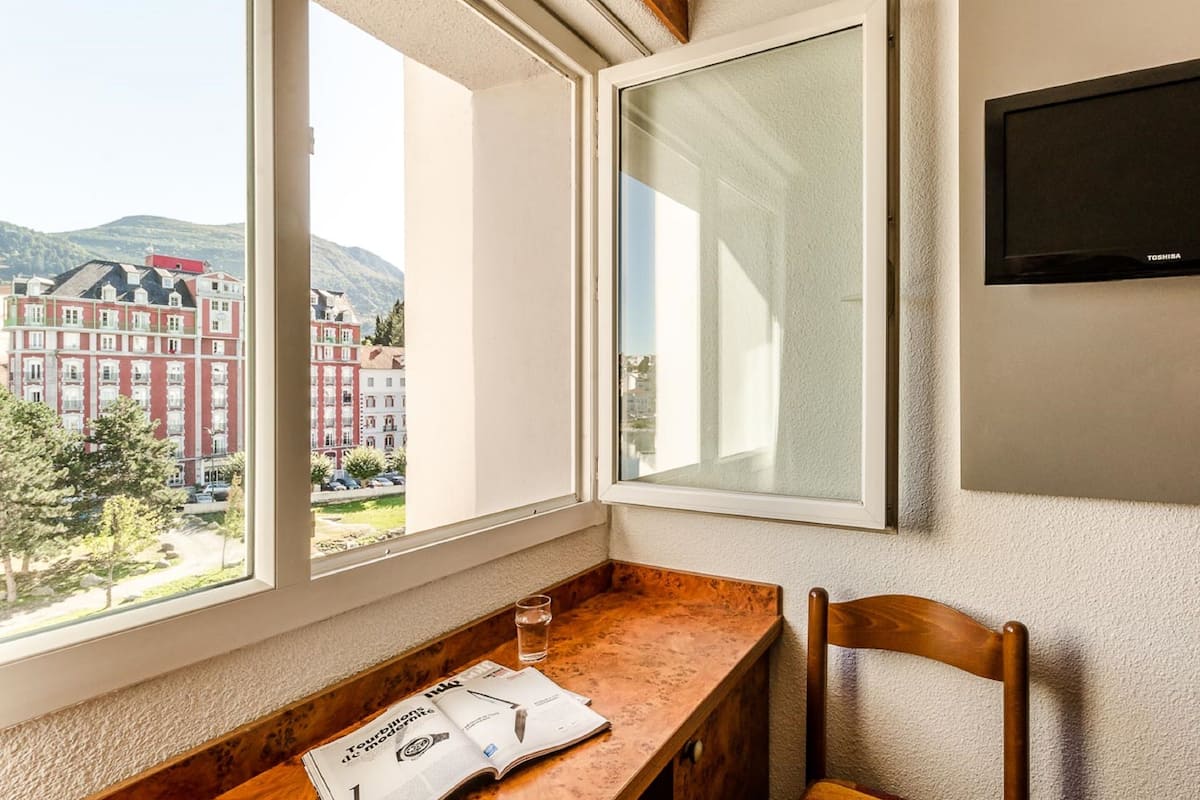 Una habitación con un escritorio de madera, una silla, un ventanal con vistas a edificios y montañas, y un televisor de pantalla plana montado en la pared.