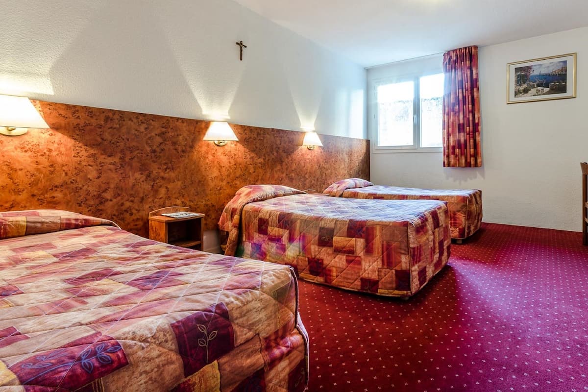 Habitación de hotel con tres camas, cabecero de madera, una cruz en la pared y una ventana con cortinas.