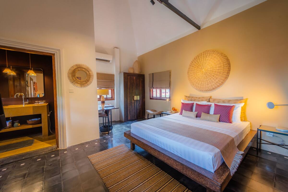 A spacious bedroom with a large bed, a wooden bench, and a bathroom with a mirror and sink.