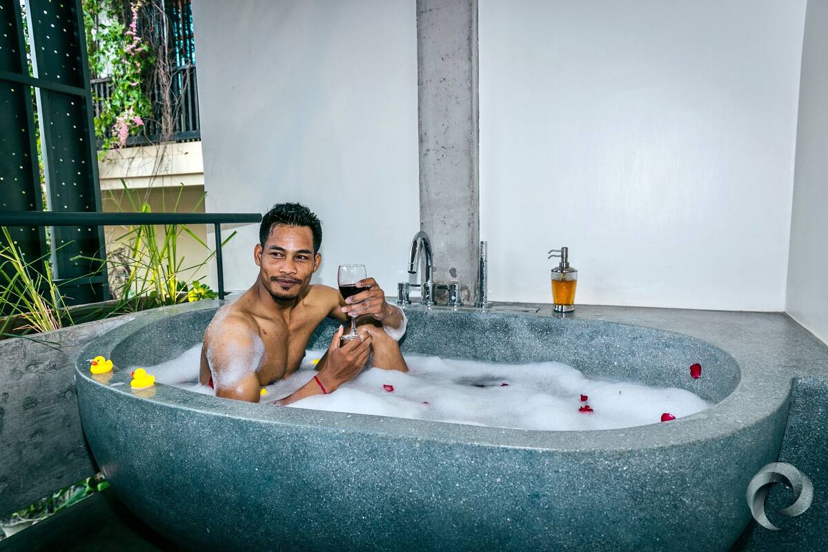 A man in a stone bathtub filled with bubbles and rose petals, holding a glass of red wine.