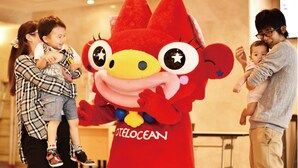 A mascot character with a red body and large eyes, standing in a room with a plain wall and a door in the background.