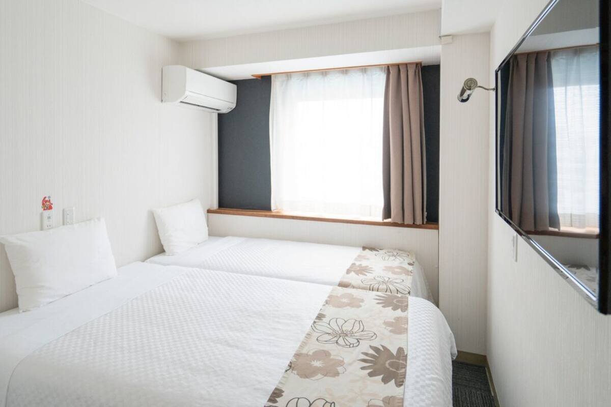 Une chambre d’hôtel avec deux lits, une grande fenêtre avec des rideaux, un climatiseur et un téléviseur à écran plat fixé au mur.