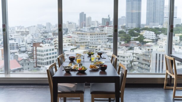 A dining table set with various dishes and drinks, positioned in front of large windows offering a cityscape view.