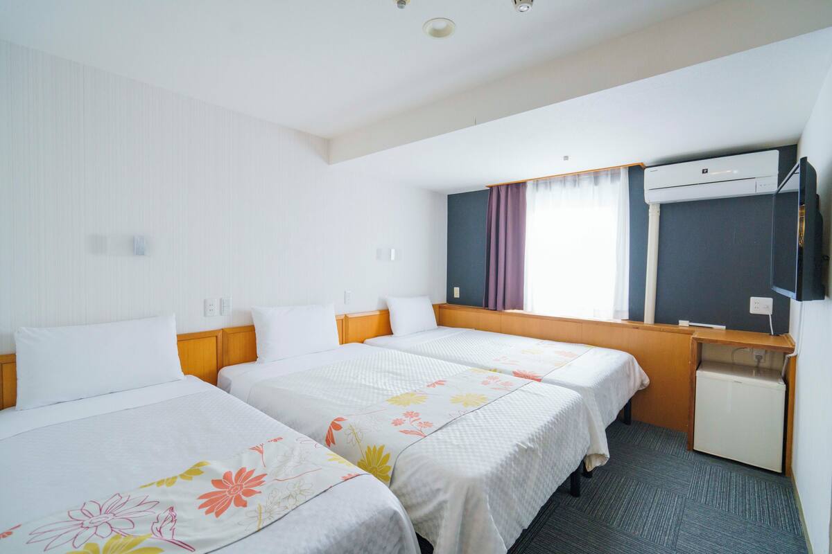 A hotel room with two beds, a desk, a small refrigerator, an air conditioning unit, and a window with curtains.