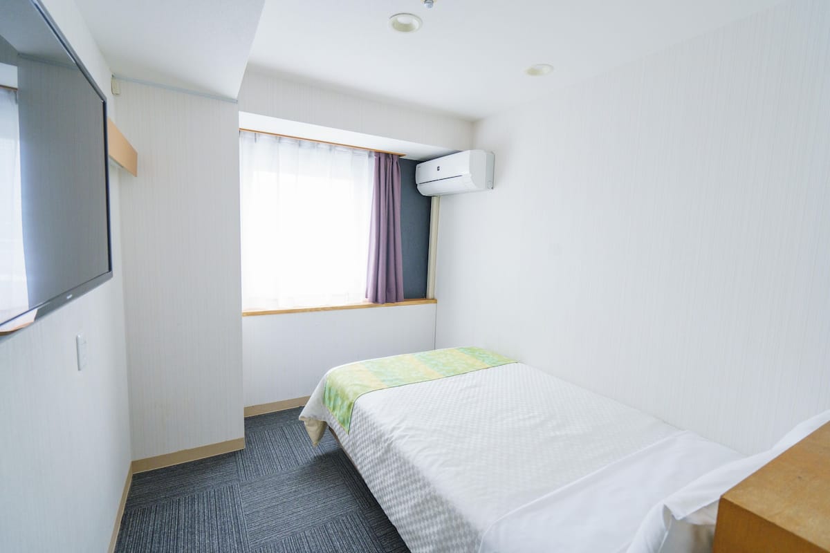 A small, neatly arranged room with a single bed, a window with curtains, an air conditioning unit, and a flat-screen TV mounted on the wall.