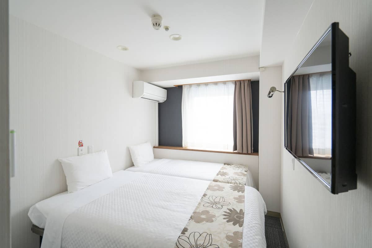 A hotel room with two beds, a large window with curtains, an air conditioning unit, and a mounted television.