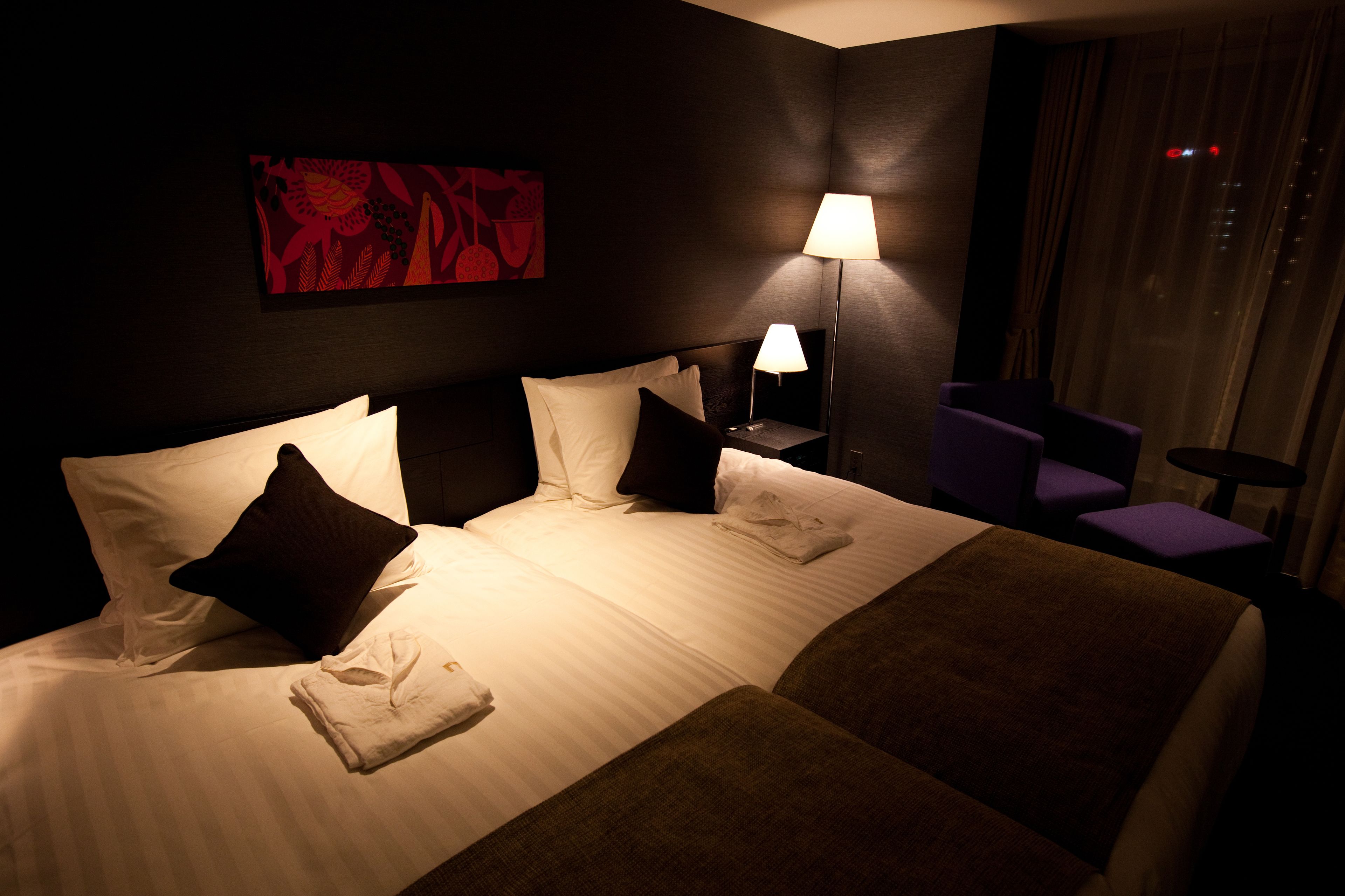 A hotel room with a large bed, a nightstand with a lamp, a chair, and a window with curtains.