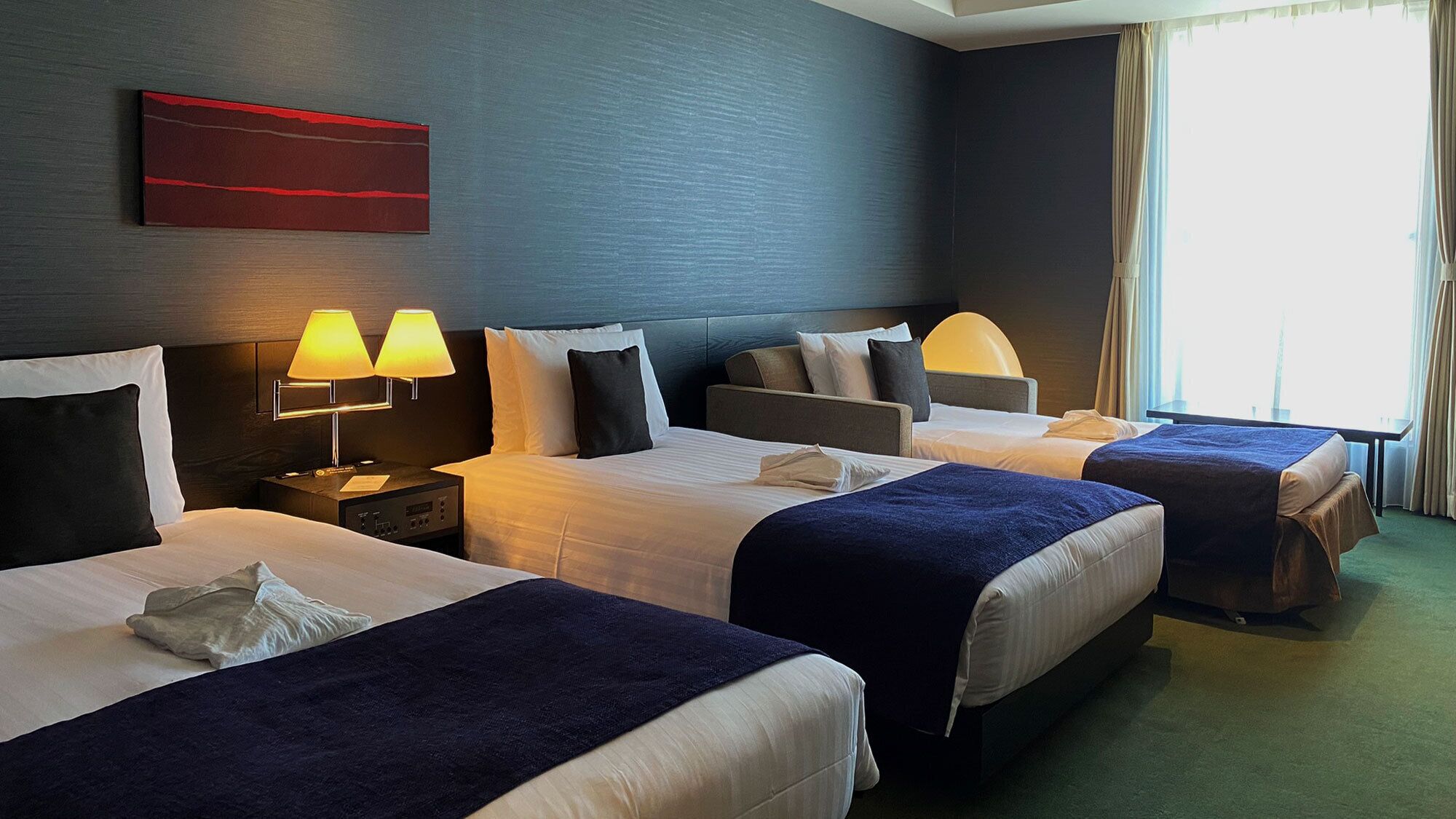 Ein Hotelzimmer mit drei Betten, einem Nachttisch mit Lampen, einer roten Akzentwand und einem großen Fenster mit Vorhängen.