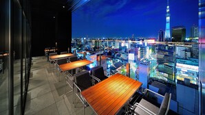 Une chambre d’hôtel située dans un immeuble de grande hauteur, offrant une vue sur la ville, et dotée d’un balcon avec tables et chaises, ainsi que d’une grande fenêtre donnant sur le paysage urbain.