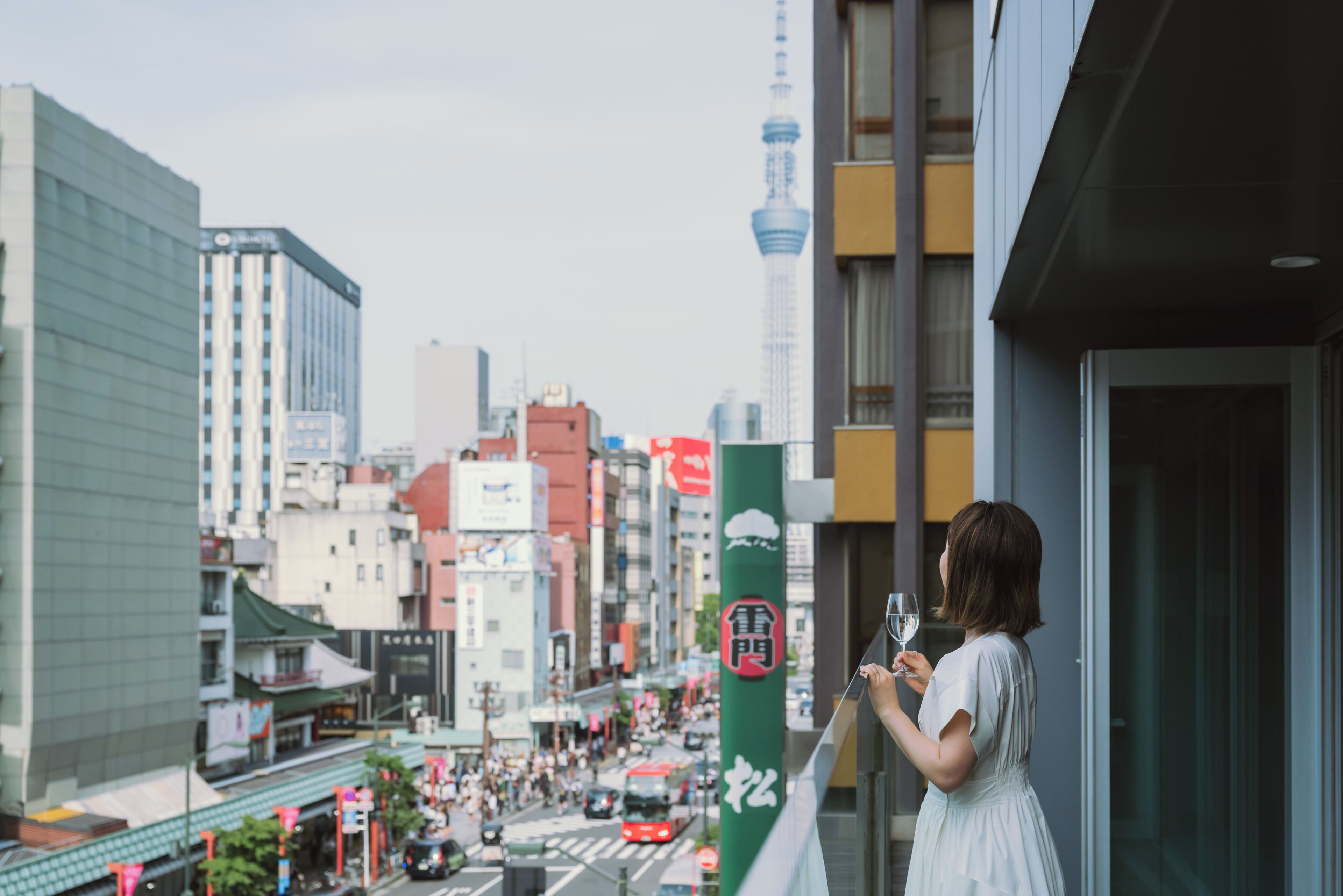 Una mujer en un balcón con vista a una concurrida calle de la ciudad, con edificios altos y una torre destacada al fondo.