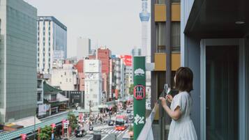 Una mujer en un balcón con vista a una concurrida calle de la ciudad, con edificios altos y una torre destacada al fondo.