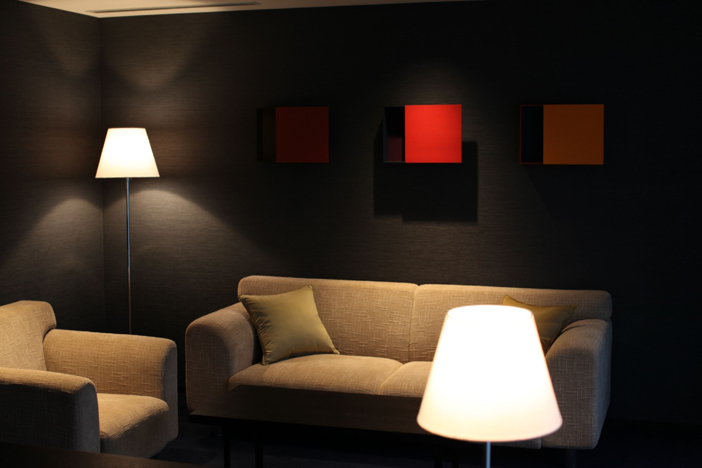 A dimly lit room with a sofa and two armchairs, a floor lamp, and three wall-mounted square light panels.