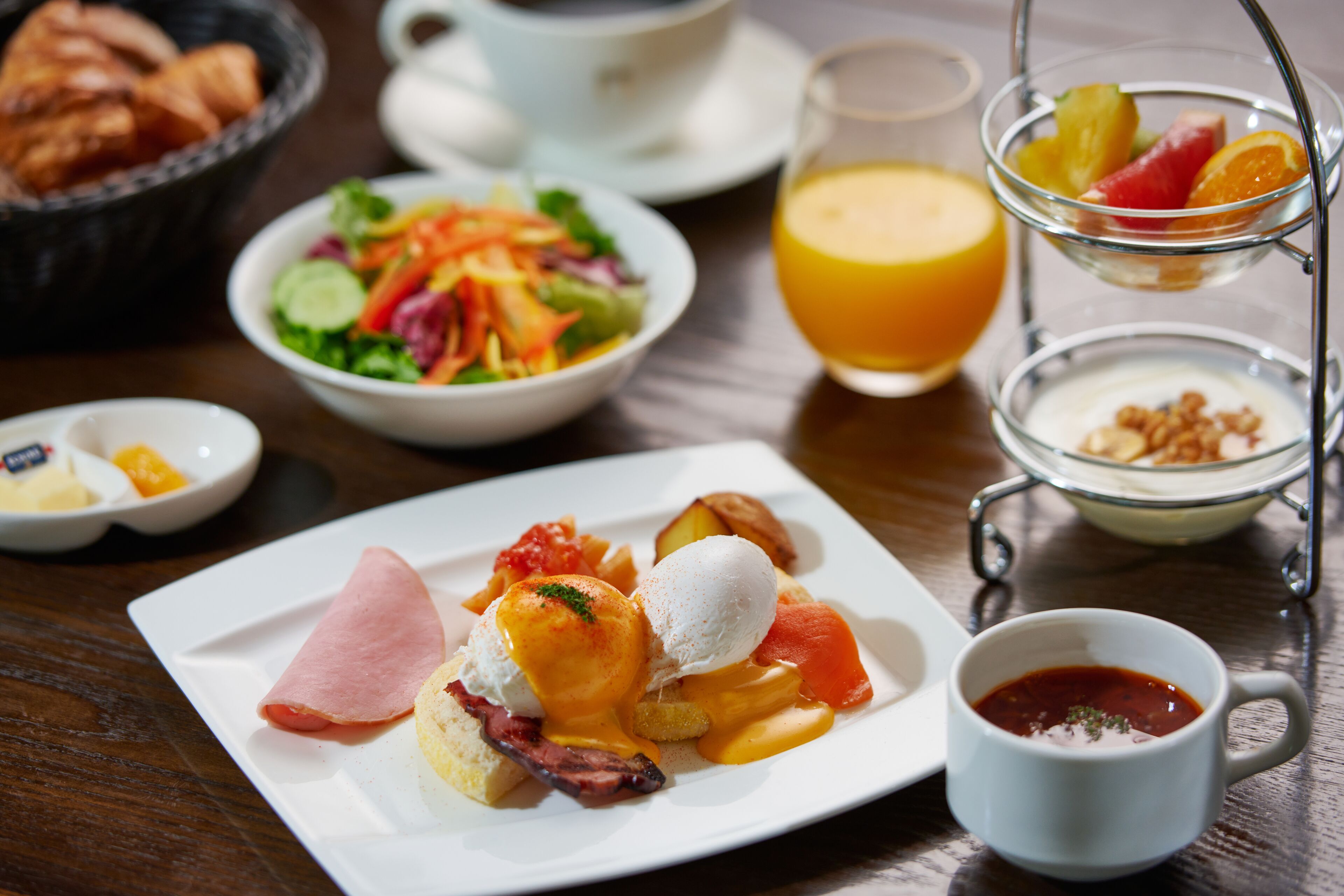 A breakfast setting with a plate of eggs benedict, a fruit bowl, a glass of orange juice, and a cup of coffee.