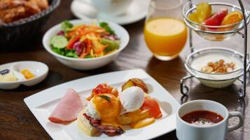 A breakfast setting with a plate of eggs benedict, a fruit bowl, a glass of orange juice, and a cup of coffee.