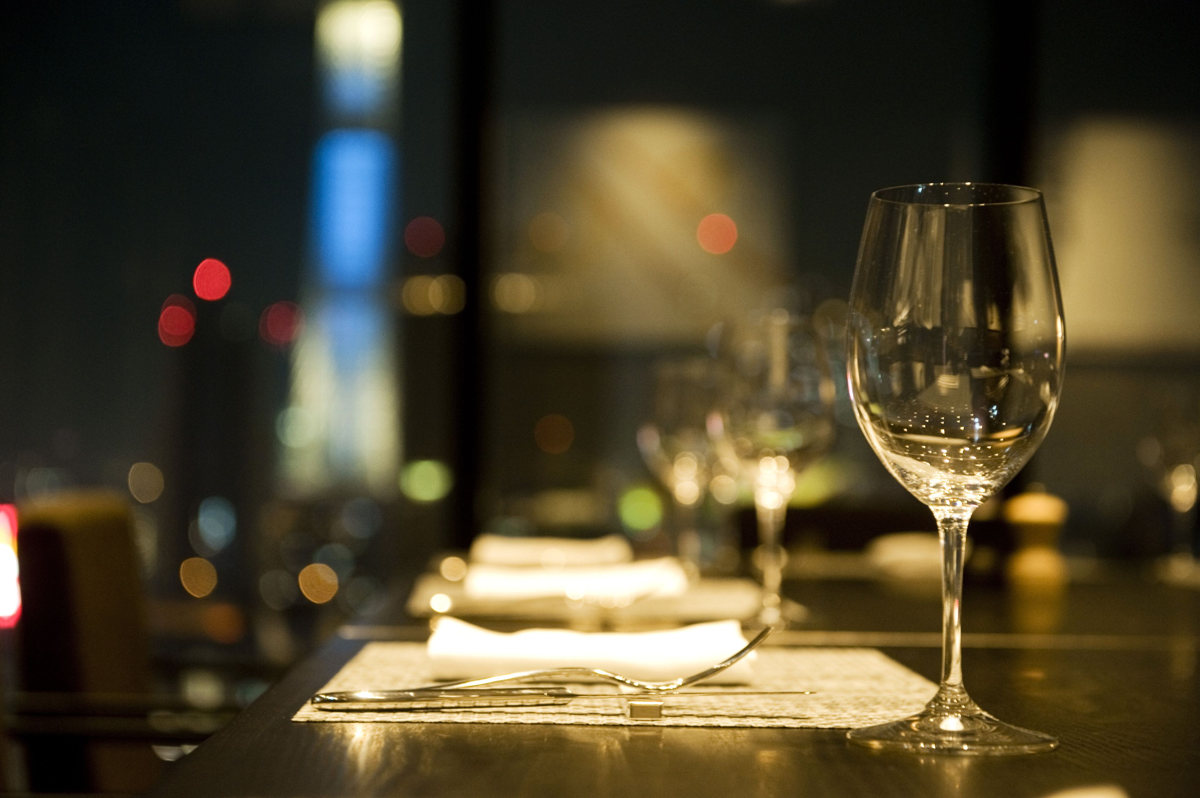 A table with a wine glass, a fork, and a napkin, with a blurred cityscape in the background.