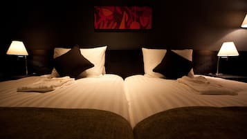 Een hotelkamer met twee bedden, elk voorzien van witte lakens en donkere kussens, met nachtkastjes en lampen ernaast. Daarboven hangt een abstract schilderij in rood.