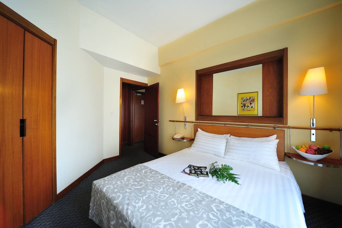 A hotel room with a large bed, a wooden wardrobe, a bedside table with a lamp, and a fruit bowl.