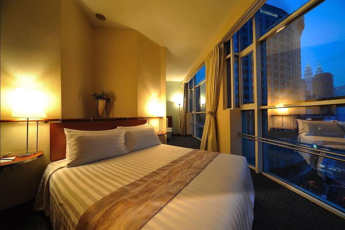 A hotel room with a large bed, a nightstand, a lamp, and a window with a city view.