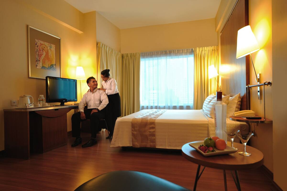 A hotel room with a bed, desk, television, and a table with fruit and a glass.