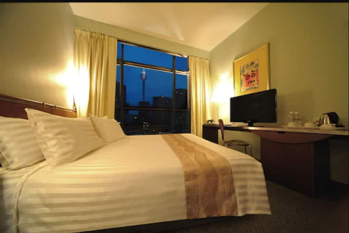 A hotel room with a large bed, a desk with a computer, and a window with a cityscape view.