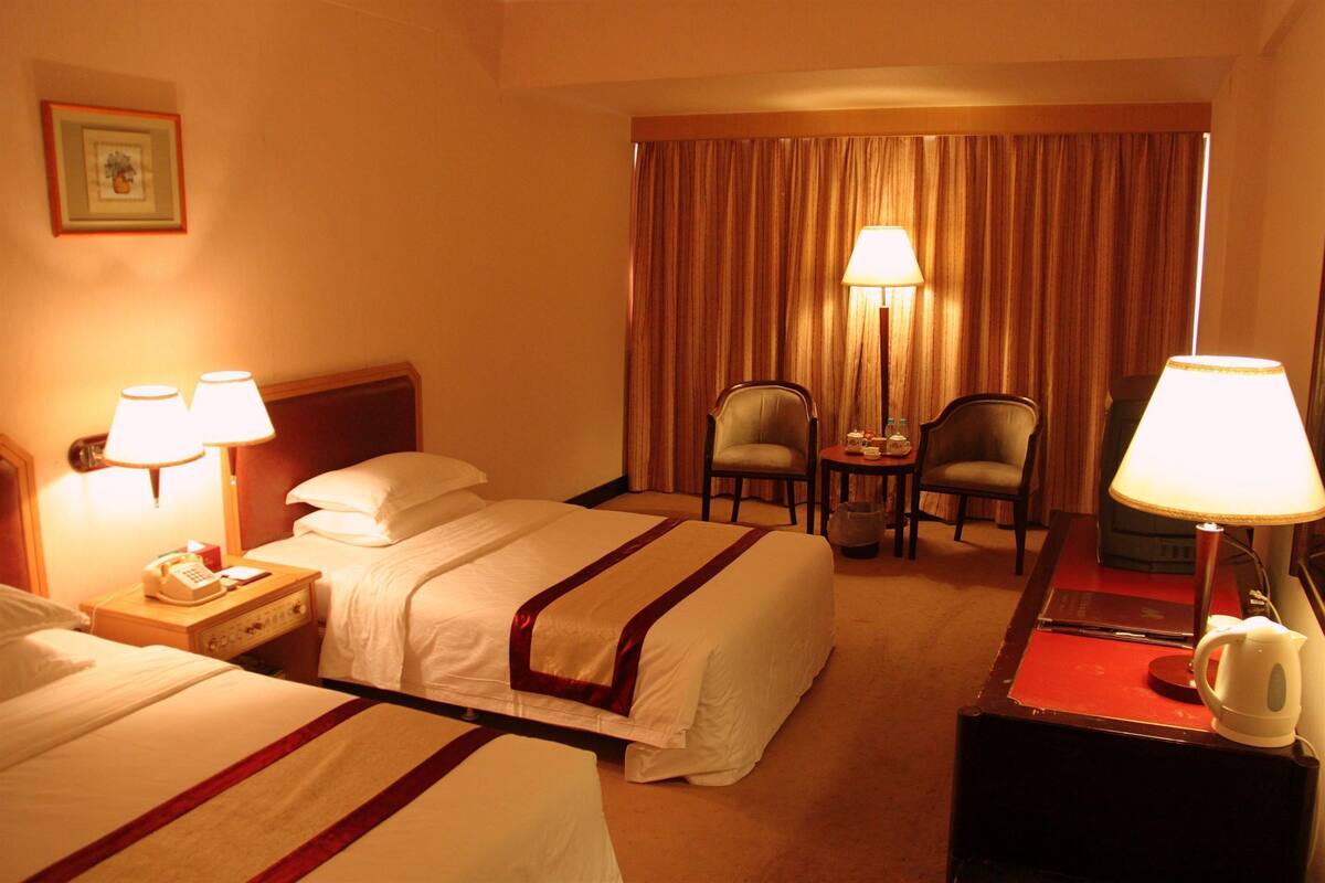 Une chambre d’hôtel avec deux lits, un bureau avec un téléphone, deux chaises, une table avec une lampe et une grande fenêtre avec des rideaux.