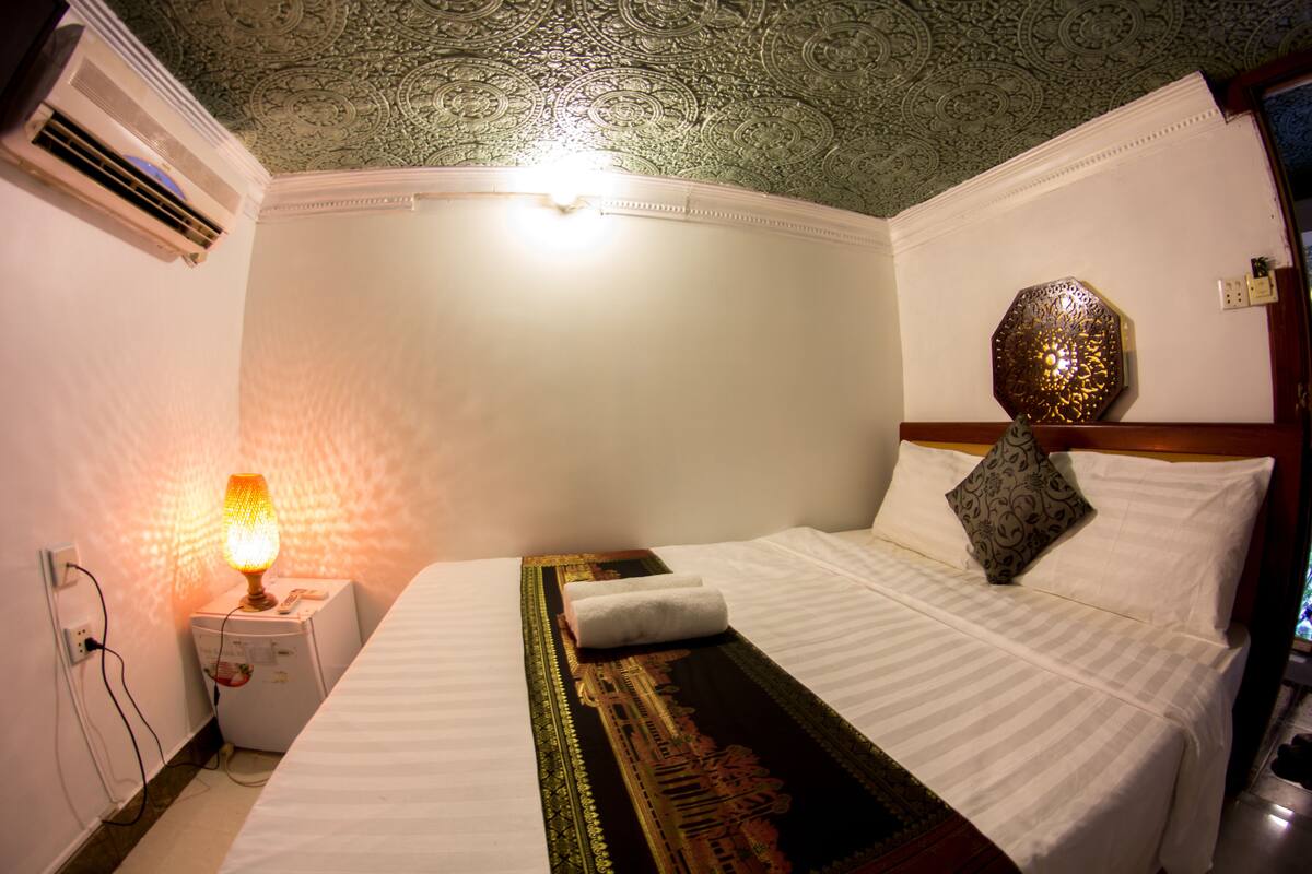 A hotel room with a bed, a nightstand with a lamp, a decorative wall piece, and an air conditioning unit.