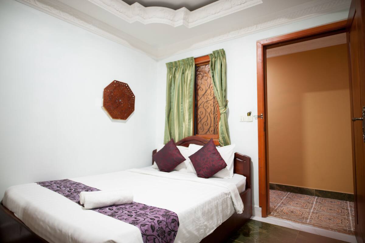 A neatly made bed with a white and purple bedspread, a wooden headboard, and two decorative pillows. A green curtain partially covers a window, and a wooden door is visible to the right.