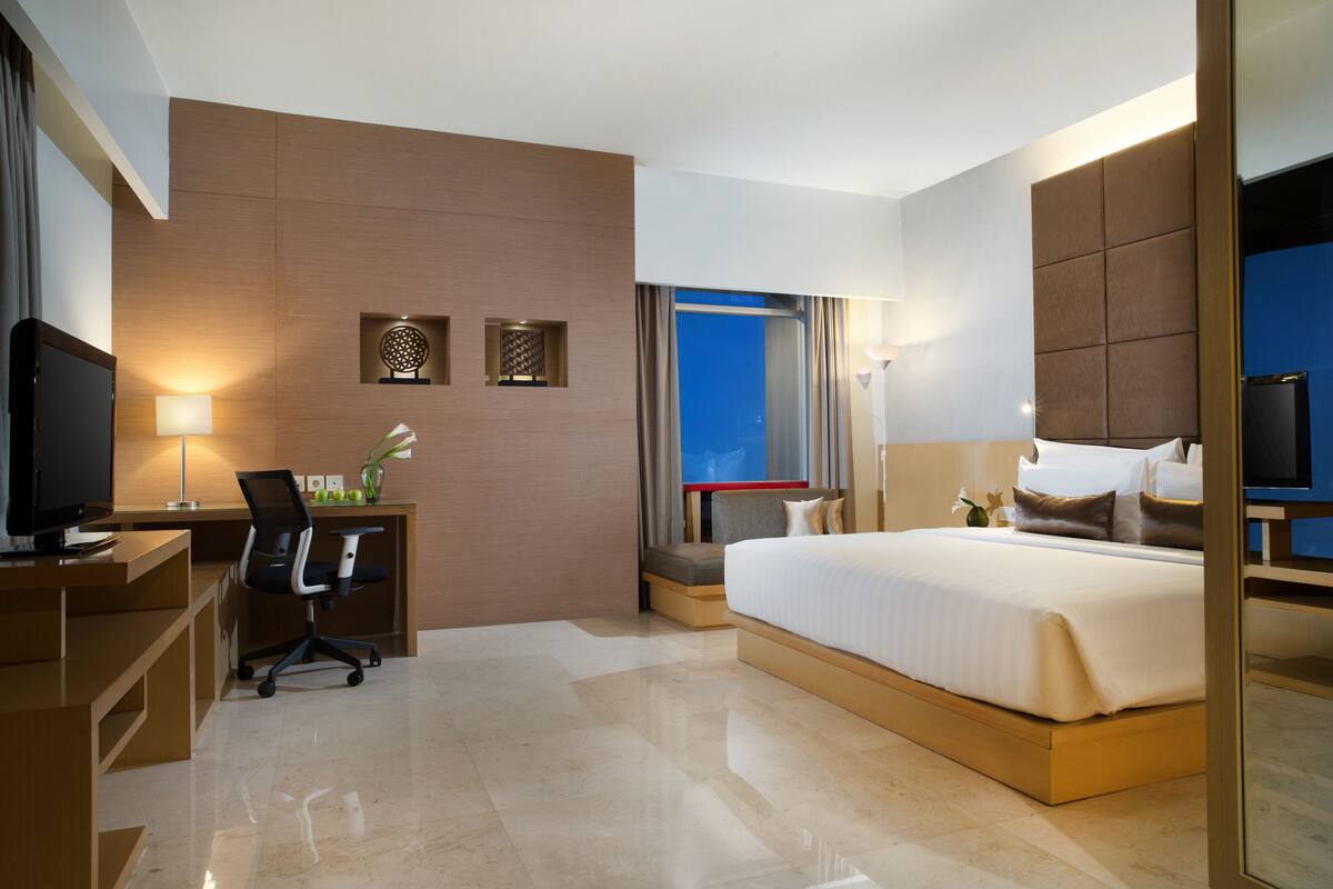 A modern hotel room with a large bed, a desk with a chair, a television, and a window with a view of the city.