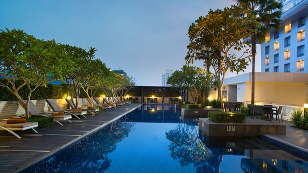 Una piscina con sillones de descanso y mesas, rodeada de árboles y un edificio con ventanas iluminadas.