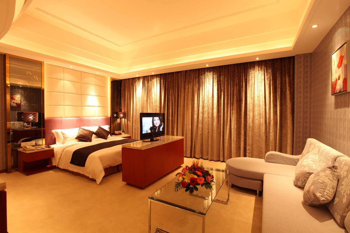 A hotel room with a bed, a sofa, a glass coffee table with flowers, a TV, and curtains.