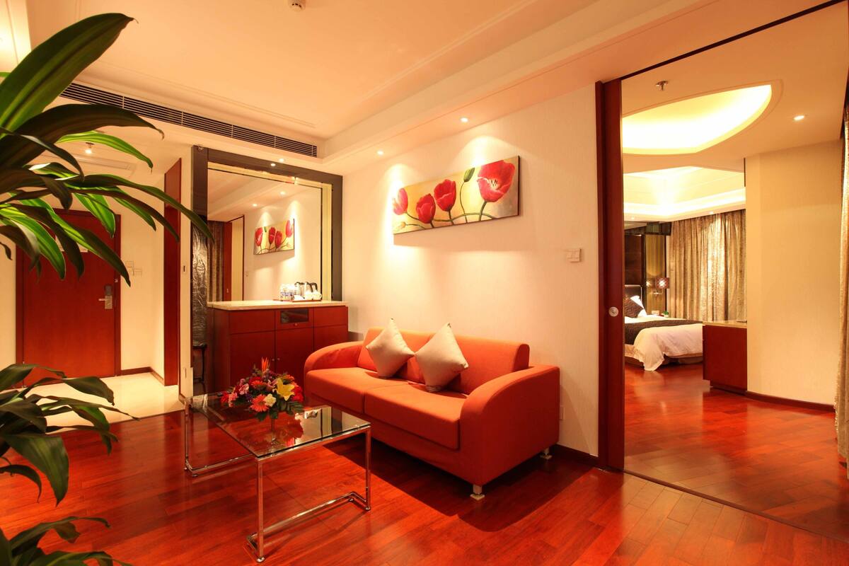 A modern hotel room with a wooden floor, a glass coffee table, a sofa with cushions, a painting on the wall, and a large mirror.