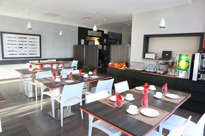 A modern dining area with multiple tables and chairs, a large menu board, and a kitchen area with appliances. - Adonis Paris Sud (Chevilly-Larue)
