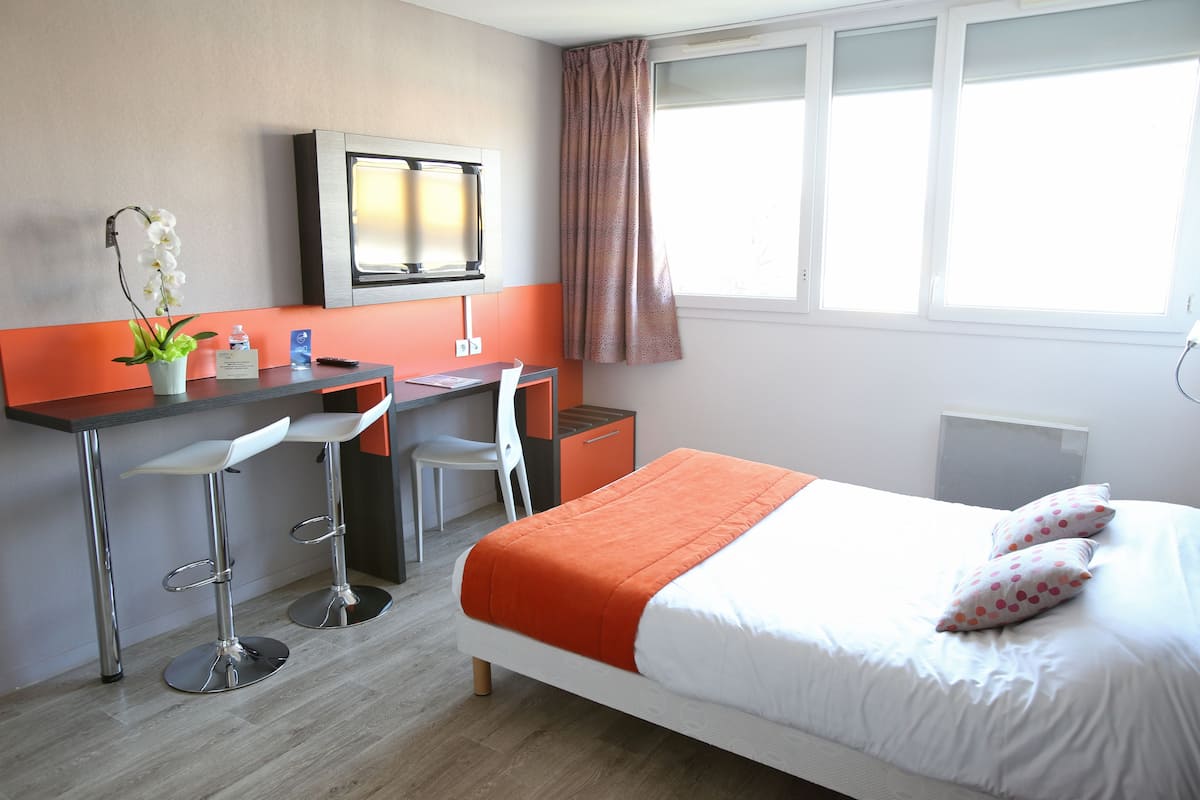 Quarto de hotel com cama, escrivaninha, dois banquinhos, televisão e janela com cortinas.